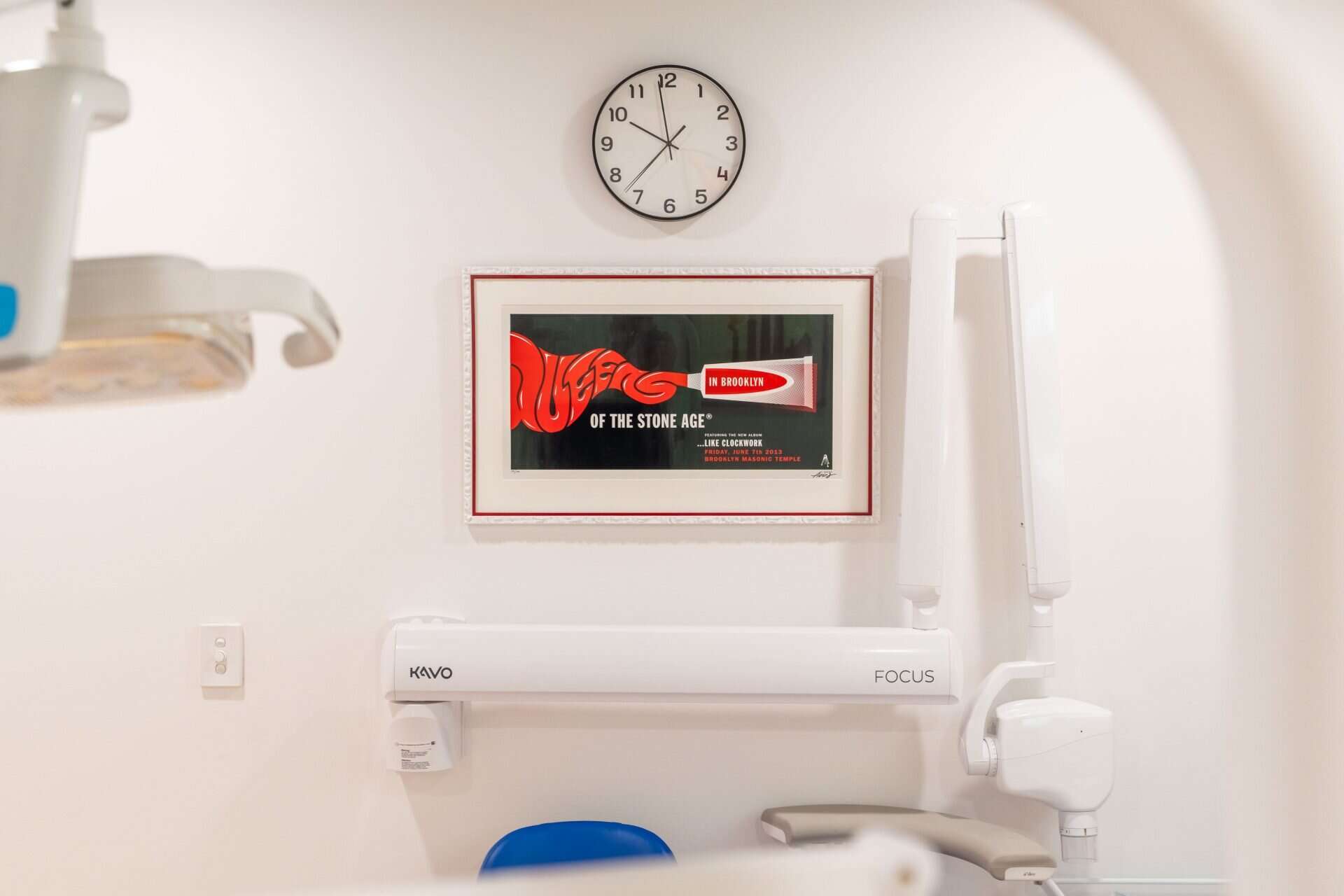 A treatment room wall, framed gig poster and wall clock above the x-ray arm beside the chair