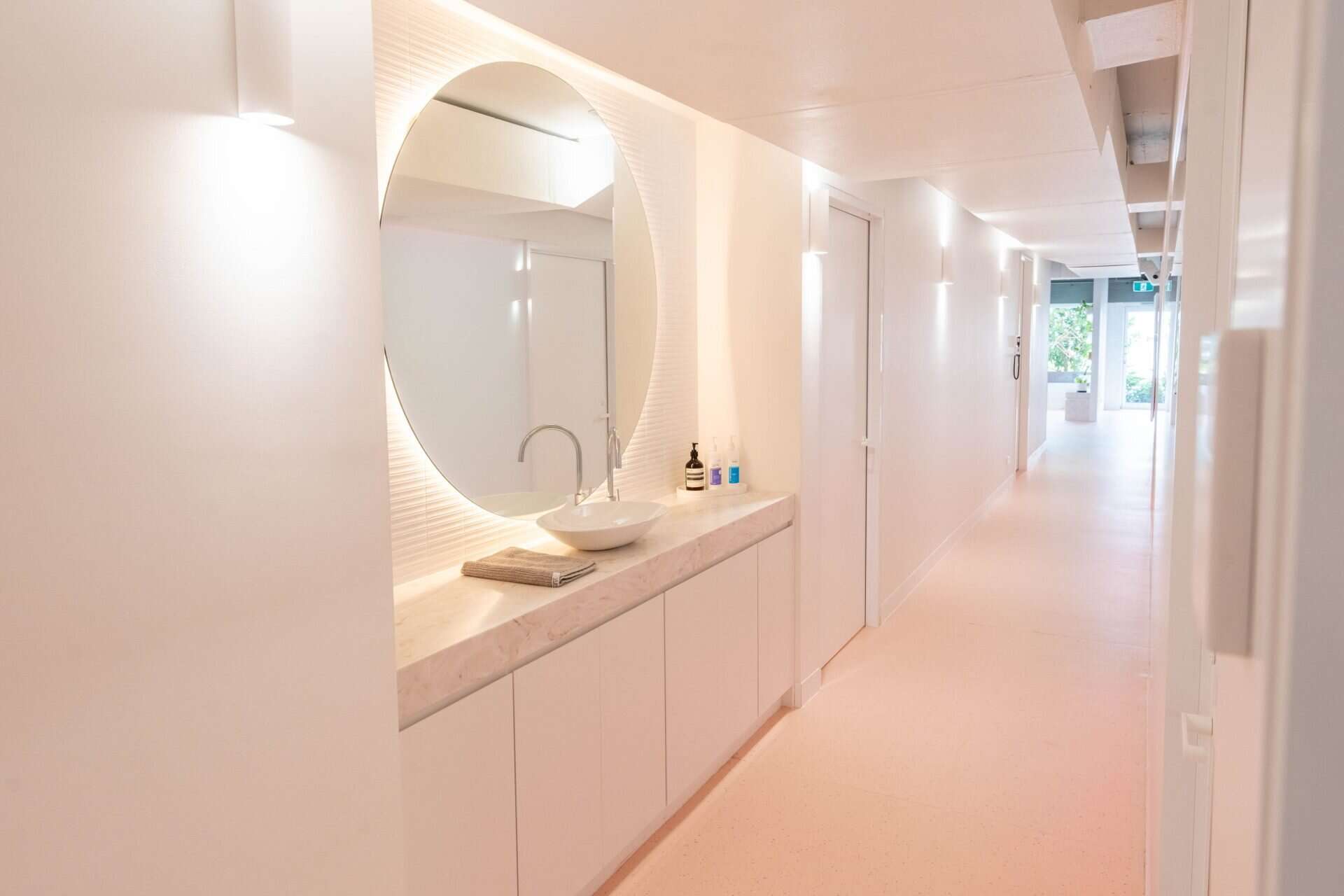 The corridor inside the practice, an oval backlit mirror over a marble vanity, with daylight and greenery through the glazed entry at the far end