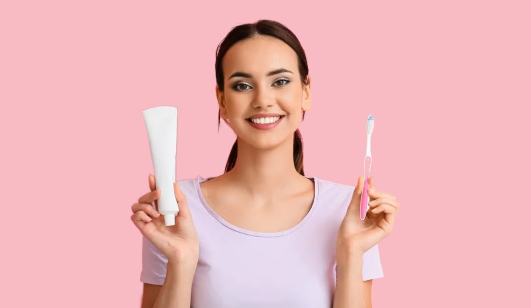 Smiling woman holding a tube of toothpaste and a toothbrush. They are wearing a pale violet tshirt and they have dark hair in a ponytail.