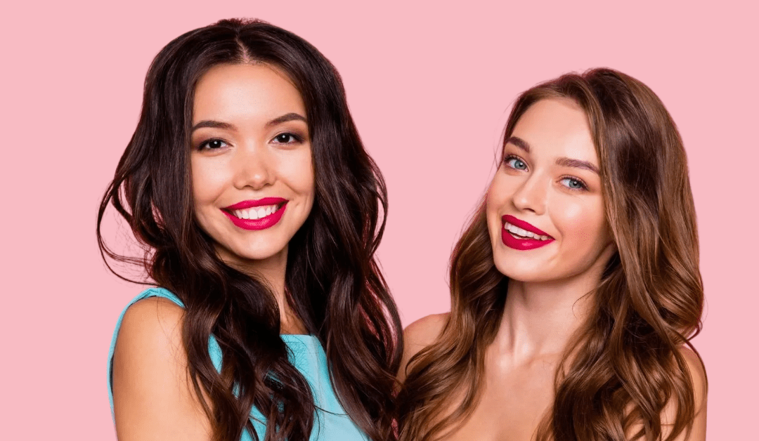 Two woman smiling widely and looking at the camera wearing red lipstick and bright outfits. One person has long dark hair and the other has long lighter hair.