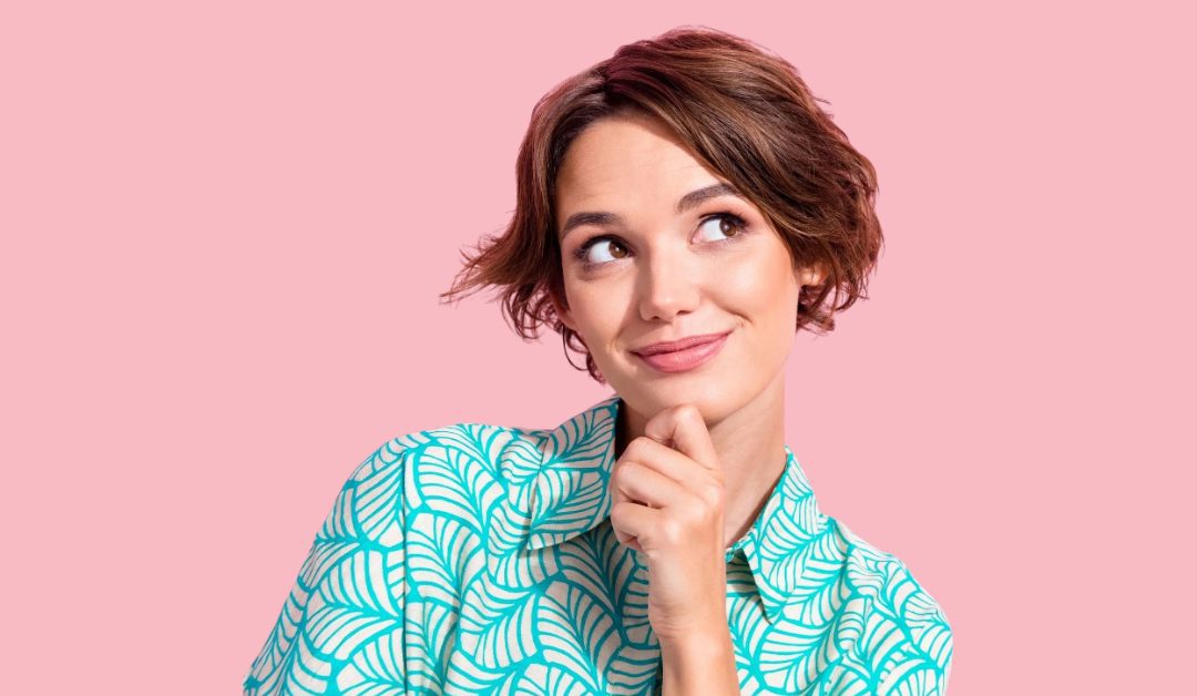 Woman with short brown hair looking to the right with her hand against her chin and a slight smile on her face. They are wearing a bright patterned aqua shirt.